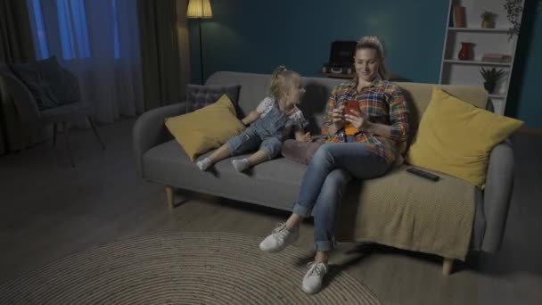 一个小女孩看电视 舒服地躺在垫子上 而她的妈妈却沉浸在智能手机里 不注意她 妈妈和女儿坐在客厅的沙发上 — 图库视频影像