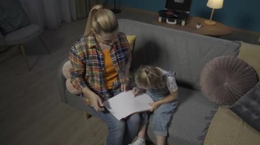 Mutlu anne akşamları oturma odasındaki kanepede otururken kızının renkli kalemlerle çizmesine yardım eder. Eğitimsel hobi, çocukların yaratıcı becerilerini geliştirir HDR BT2020 HLG Materyal