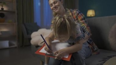 Mutlu anne akşamları oturma odasındaki kanepede otururken kızının renkli kalemlerle çizmesine yardım eder. Anneler kucağında oturan ve Noel ağacı çizen bir kız. Eğitici eğlence.