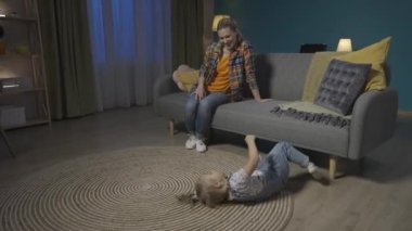 Anne ve küçük kızı akşamları oturma odasında eğleniyorlar. Küçük aktif kız yerde sallanıyor ve anne koltukta oturmuş HDR BT2020 HLG materyallerini izliyor ve gülüyor.