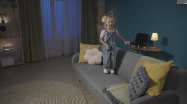 Oturma odasındaki kanepede zıplayan küçük kız, gülüyor ve ellerini çırpıyor. Çocuk oyunları ve evde eğlence. Hiperaktivite konsepti HDR BT2020 HLG Malzeme