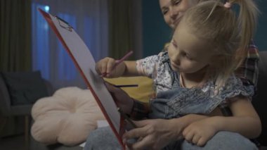 Mutlu anne kızının oturma odasındaki kanepede otururken renkli kalemlerle resim yapmasına yardım ediyor. Kızı annelerinin kucağına oturur ve annesi de elinde çocuk için kağıt olan bir pano tutar.