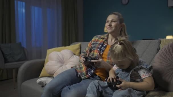 ママと娘はジョイスティックでビデオゲームをしている ママと娘は一緒に夜を過ごし テレビの前にソファーのリビングルームに座って楽しんでいました 家族の親の日 — ストック動画