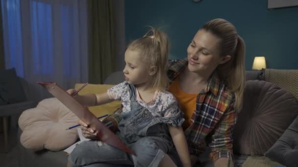 Onnellinen Äiti Auttaa Tytärtään Piirtämään Värillisillä Kynillä Istuessaan Olohuoneen Sohvalla — kuvapankkivideo