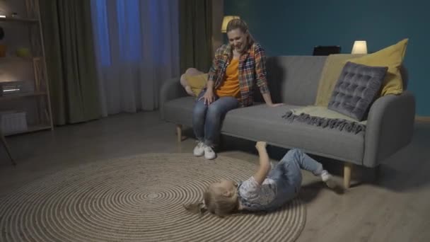 ママと小さな娘は夜のリビングで楽しんでいました 小さなアクティブな女の子が床に振りかけていて お母さんはソファーで見ていて笑っています Hdr Bt2020 Hlg — ストック動画