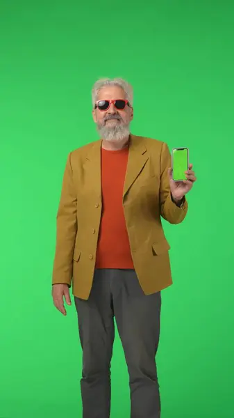 Creative modern seniors concept. Portrait of senior stylish hipster on Chroma key green screen background, elegant man holding smartphone at camera. Advertising area, workspace mockup. Vertical photo.