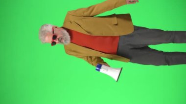 Yaratıcı yaşlılar konsepti. Şık ceketli ve gözlüklü yaşlı hippi portresi Krom anahtar yeşil ekranda, megafonla bağırıyor. Reklam alanı, çalışma alanı modeli. Dikey video.