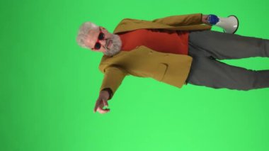 Yaratıcı yaşlılar konsepti. Şık ceketli ve gözlüklü yaşlı hippi portresi Krom anahtar yeşil ekranda megafonla konuşuyor. Reklam alanı, çalışma alanı modeli. Dikey video.
