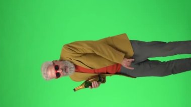 Yaratıcı yaşlılar konsepti. Şık ceketli, gözlüklü, krom renkli yeşil ekranda şampanya içen kıdemli hippi portresi. Reklam alanı, çalışma alanı modeli. Dikey video.