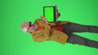 Yaratıcı yaşlılar konsepti. Şık ceketli ve gözlüklü yaşlı hippi adamın portresi. Dizüstü bilgisayar, anahtar yeşil ekran, kameraya poz veriyor. - Reklam alanı, çalışma alanı modeli. Dikey video.