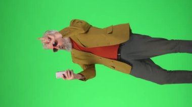 Yaratıcı yaşlılar konsepti. Şık ceketli ve gözlüklü yaşlı hippi portresi Krom anahtar yeşil ekranda, akıllı telefondan selfie çekiyor. Reklam alanı, çalışma alanı modeli. Dikey video.