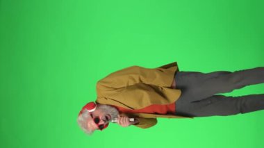 Yaratıcı yaşlılar konsepti. Ceketli kıdemli hippi adam ve Chroma 'da gözlüklü yeşil ekran, kulaklıklı, karaoke mikrofonla dans eden. Reklam alanı, çalışma alanı modeli. Dikey video.