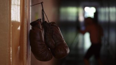 Atış, kahverengi boks eldivenlerinin asıldığı paslı çivili kapı pervazını gösteriyor. Arka planda boks eldivenli bir adam bir rafta durur ve dövüşten önce yumruklarını biler.