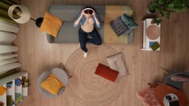 Yukarıdaki karede, hamile bir kadın bir odada kanepede oturuyor. Kablosuz kulaklıklarını takmış ve müzik dinliyor, hoşuna gidiyor. O zor bir zaman geçiriyor bu yüzden dinleniyor. Üst görünüm.
