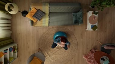 Yukarıdaki karede, hamile bir kadın bir odada lastik bir fitness topunun üzerinde mavi bir şekilde oturuyor. Karnını okşuyor, sonra gerilmek için kollarını kaldırıyor. Bitirdikten sonra, oturup karnını okşamaya devam ediyor..