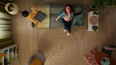 Yukarıdan çekimde, hamile bir kadın bir odada kanepede oturuyor. Gözlerini kapayıp karnına sarılıyor, acı içinde kıvranıyor, çok kötü hissediyor. Bir bebek bekliyor. Üst görünüm.