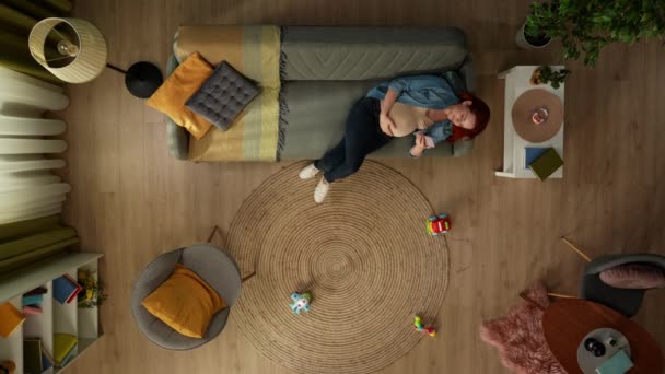 在上面的镜头中 一个孕妇躺在房间的沙发上 她拿着电话 看着里面的东西 她是快乐和休息的 附近的地板上有儿童玩具 — 图库视频影像