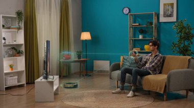 Akıllı ev ve modern yaşam tarzı reklam konsepti. Oturma odasındaki kanepede oturan genç adam elinde akıllı telefonuyla hoparlörü açıyor ve robot elektrikli süpürgeyle bilgi paylaşımı yapıyor..