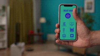 Akıllı ev yaratıcı reklam konsepti. Erkek cep telefonunun yakın çekimi. Adam ışıkları açıyor, robot temizleyici, akıllı telefon uygulaması ile perdeleri açıyor, arayüz simgeleri ile ekrana odaklanıyor.