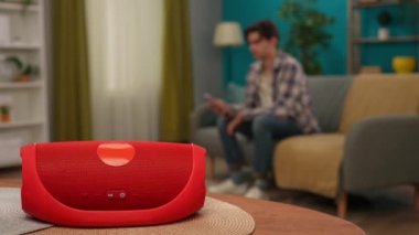 Akıllı ev ve modern teknoloji yaratıcı reklam konsepti. Kırmızı müzik hoparlörünü kapat. Kanepede oturan adam akıllı telefonu tutuyor ve telefon uygulamasını kullanarak hoparlörü açıyor..