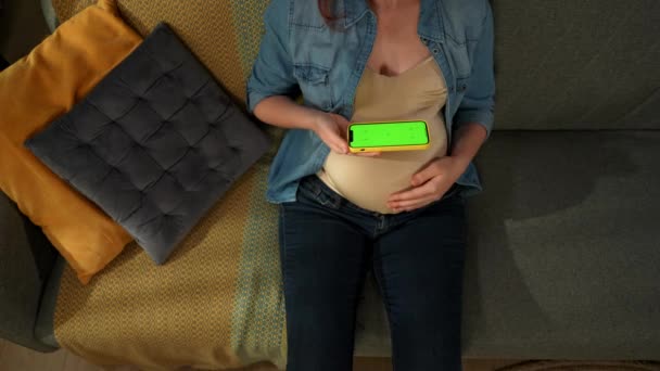近照显示一个孕妇坐在沙发上 她手里拿着一个有绿色屏风的电话 她检查他 这是你的广告 顶部视图 — 图库视频影像