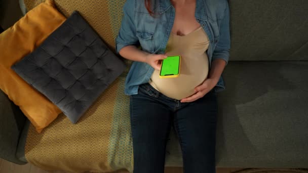 クローズアップショットは ソファーに座っている妊婦を示しています 彼女は彼女の手に緑色のスクリーンを持った携帯電話を持っている 彼女は彼を調べている ここにあなたの広告があるかもしれません トップビュー ミディアムショット — ストック動画