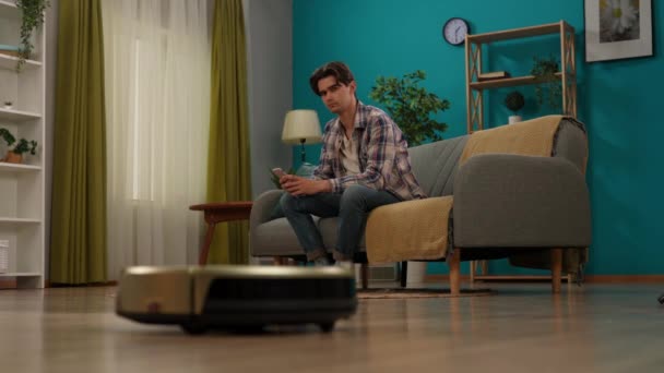 スマートハウスとモダンテクノロジーのクリエイティブ広告コンセプト ソファーに座ってスマートフォンを保持している男性は アプリを使用してロボット掃除機をオンにし バッテリーのインフォグラフィックはクリーナー上に見えます — ストック動画