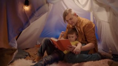 Aile zamanı konsepti. Oğluyla birlikte oturma odasındaki çadırda kitap okuyan bir baba. Oğlu sırt üstü babasının göğsüne yaslanmış yatıyordu ve babası kitabı işaret ettiğinde baş parmağını kaldırdı.