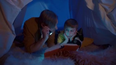 Aile zamanı konsepti. Baba ve oğul karanlık bir oturma odasındaki çadırda kitap okuyorlar. Bir adam ve bir çocuk, yumuşak kürklü bir ekose üzerinde göbeklerinin üzerine uzanır. Adam, el fenerini kitaba doğru tutar. Oğlu da...