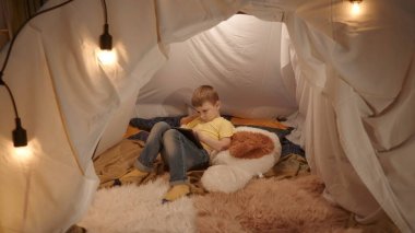 Çocuk elinde tabletle büyük yumuşak bir oyuncağın üzerinde yatıyor. Küçük bir çocuk geceyi, oturma odasında kurulmuş bir çadırda, bir tablette online oyun oynayarak geçirir.