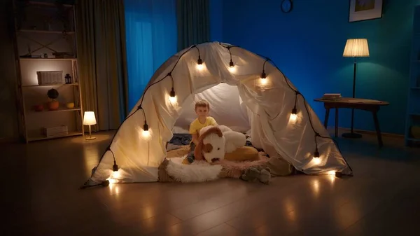 Portrait of a small boy, with a large soft toy in the form of a dog in a tent decorated with garlands. Boy with soft toy sitting on fur plaids in a tent. Concept of happy childhood