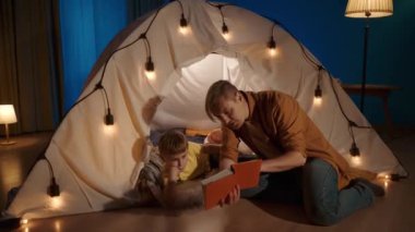 Aile zamanı konsepti. Bir baba oğluna kitap okuyor, sayfalarda bir şeyler anlatıyor ve gösteriyor, çocukça sorulara olumlu cevap veriyor. Oturma odasında bir adam oturuyor.