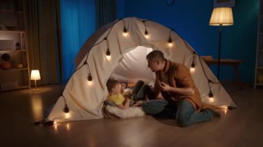 Baba ve oğlunun kurabiye yerken ve baş parmağını kaldırırken, akşam atıştırması. Oturma odasında çelengin yanında oturan bir adam içinde bir çocuk olan çadır ve bir tabak kurabiyeyle.