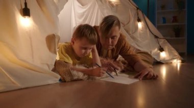 Baba ve oğul renkli kalemlerle bir deftere çiziyorlar. Bir adam çocuğa yardım ediyor. Baba ve oğul, oturma odasındaki çelenk çadırında göbeklerinin üstünde yatıyorlar.