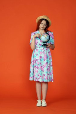 Seyahat ve tatil reklamları konsepti. Kadın modelin stüdyo fotoğrafı. Küçük bir küre tutan kadın, üzgün surat ifadesi, haritanın hedefini gösteren turuncu arka planda izole edilmiş bir kadın..