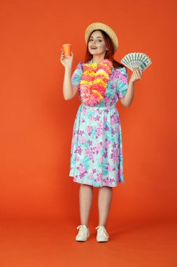 Seyahat ve tatil reklamları konsepti. Kadın modelin stüdyo fotoğrafı. Kağıt bardakta çiçekli bir kadın ve banknotların hayranı, kameraya bakıp gülümsüyor, turuncu arka planda soyutlanmış..