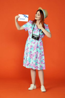 Seyahat ve tatil reklamları konsepti. Kadın modelin stüdyo fotoğrafı. Şapkalı kadın elinde hediye sertifikası ile mutlu bir şekilde gülümsüyor, gazeteye bakıyor, turuncu arka planda soyutlanmış..