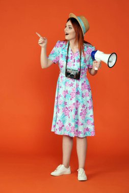 Seyahat ve tatil reklamları konsepti. Kadın modelin stüdyo fotoğrafı. Kadın şaşırtıcı bir şekilde işaret eden bir şeye bakıyor ve elinde turuncu arka planda izole edilmiş küçük bir megafon tutuyor..