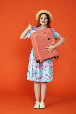 Seyahat ve tatil reklamları konsepti. Kadın modelin stüdyo fotoğrafı. Şapkalı kadın kameraya bakıyor gülümsüyor, bavulu tutuyor, başparmaklarını gösteriyor, turuncu arka planda izole edilmiş..