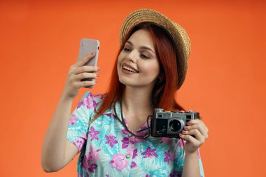Seyahat reklam konsepti. Kadın modelin stüdyo fotoğrafı. Mutlu bir şekilde gülümseyen, elinde akıllı telefon ve eski bir kamerayla selfie çeken, turuncu arka planda izole edilmiş neşeli bir kadın..