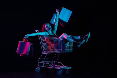 Kara Cuma ve mevsimler satış reklamları konsepti. Alışveriş arabasında oturan çekici bir kadın alışveriş torbası tutan eller, neşeli bir yüz ifadesi. Siyah arkaplanda izole edilmiş neon ışığı.