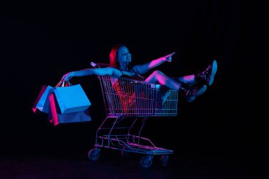Kara Cuma ve mevsimler satış reklamları konsepti. Alışveriş arabasında elinde poşetlerle oturan ve yan tarafı gösteren çekici bir kadın, mutlu bir ifade. Siyah arkaplanda izole edilmiş neon ışığı
