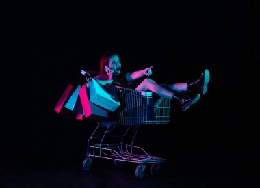 Kara Cuma ve mevsimler satış reklamları konsepti. Alışveriş arabasında oturan çekici bir kadın akıllı telefondan şaşkın bir ifadeyle işaret ediyor. Siyah arkaplanda izole edilmiş neon ışığı.