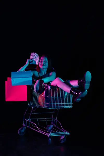 Black friday and seasons sale advertisement concept. Attractive woman sitting in shopping cart holding fan of money bills smiling face with closed eyes. Isolated on black background in neon light.