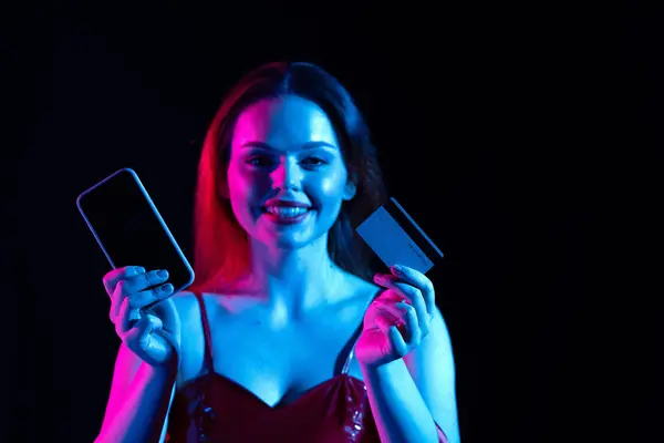 ブラックフライデーとシーズンセール広告コンセプト スマートフォンや銀行のクレジットカードを持っている魅力的な女性 カメラで笑顔 オンラインショッピングの支払い ネオンライトの黒い背景に隔離される — ストック写真