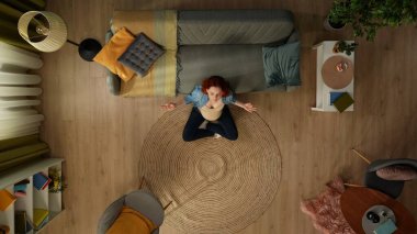 Üst katlı bacaklı karede bir kadın bir odada yerde oturuyor. Sakinliğini, huzurunu, konsantrasyonunu gösteriyor. Yakınında uzanıp meditasyon yaptığı bir kanepe var. Üst görünüm.