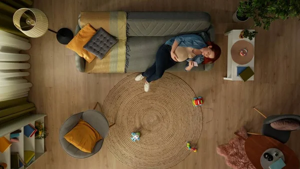 在上面的镜头中 一个孕妇躺在房间的沙发上 她拿着电话 看着里面的东西 她是快乐和休息的 附近的地板上有儿童玩具 — 图库照片