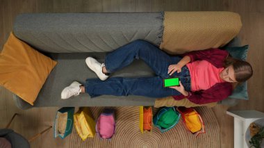 Alışveriş, eğlence ve yaşam tarzı reklam konsepti. Üst görüntü, genç bir kızın akıllı telefon anahtarı yeşil ekran ile kanepede yattığını gösteriyor. Reklam alanı çalışma alanı modellemesi.