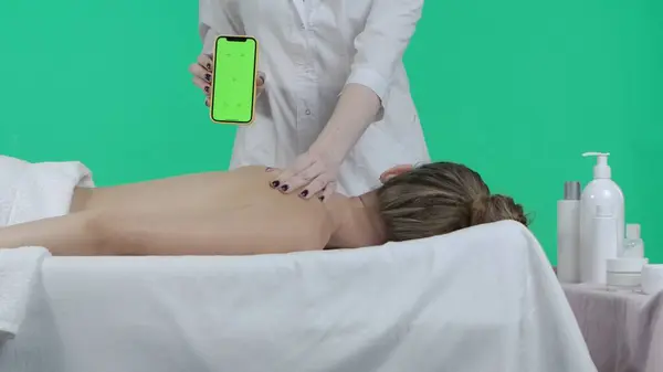 Body wellbeing creative concept. Beauty salon room, female on massage table, masseur holding smartphone with Chroma key green screen background, touching client back, advertising area workspace mockup