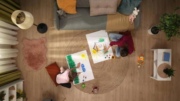 In the picture from above sit on the floor of a woman and the child they draw on the sheets of each their own. There is a sofa, on the floor are paints and toys. Theyre having fun, wondering.